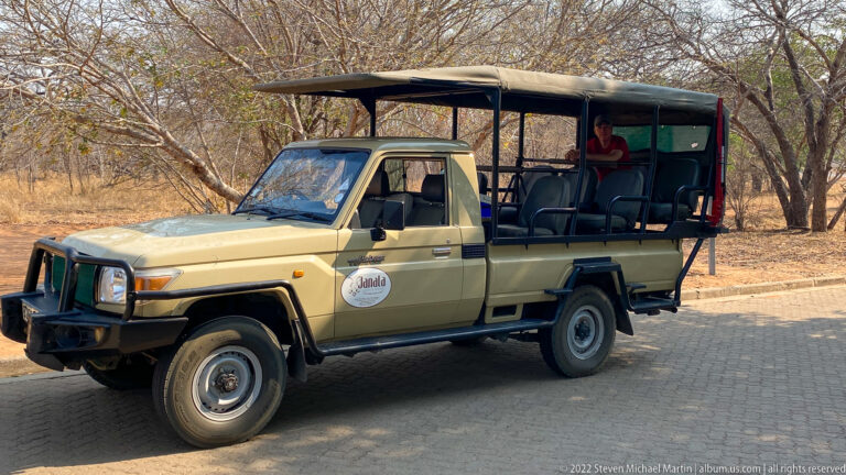 Open-sided Toyota safari vehicle with elevated passenger seats, canvas roof, bull bar, and rear step parked near dry woodland.