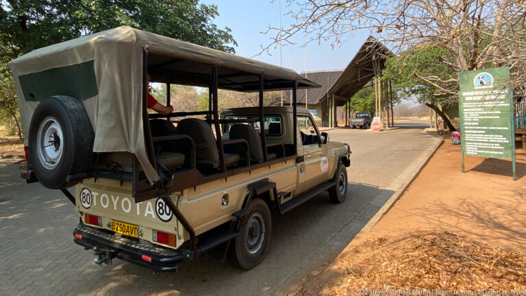 Rear view of an open safari vehicle approaching Sedudu Wildlife Gate, with the gate canopy and green park sign ahead.