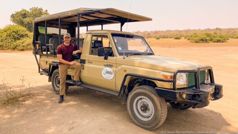 Smiling safari traveler standing beside a tan open-sided game-drive vehicle with elevated seats and canvas roof on a sandy track.
