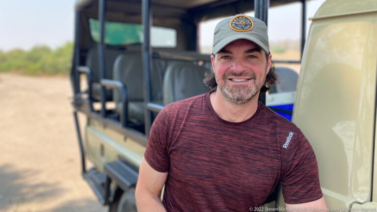 Close portrait of a smiling traveler in a cap and dark red shirt beside the open passenger area of a safari vehicle.