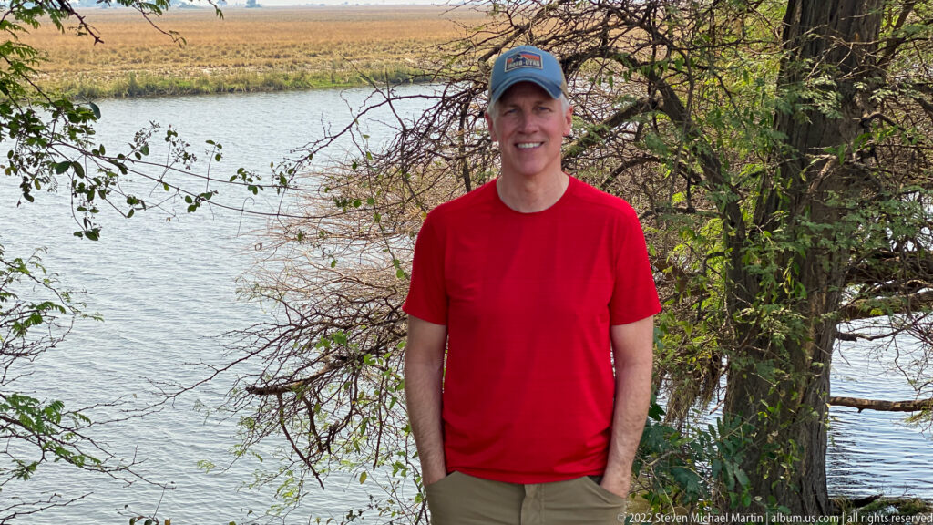 Traveler in a red shirt and cap standing beneath trees beside broad calm water in Chobe National Park.