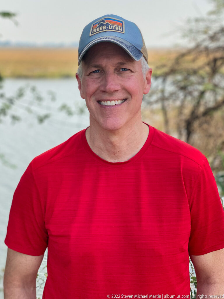 Close portrait of a smiling traveler wearing a red shirt and cap, with river water and floodplain softly blurred behind.