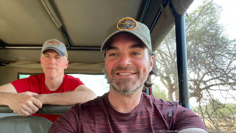 Two travelers seated inside an open safari vehicle, one close to the camera and another leaning forward from the rear bench.