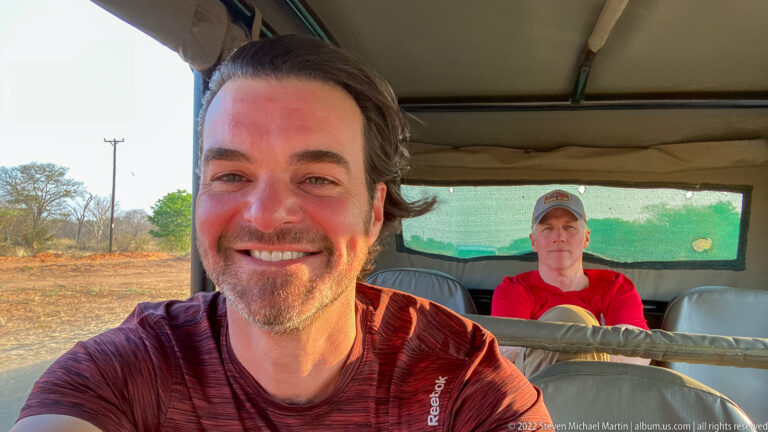 Two travelers inside an open safari vehicle, with one smiling close to the camera and another seated on the rear bench.