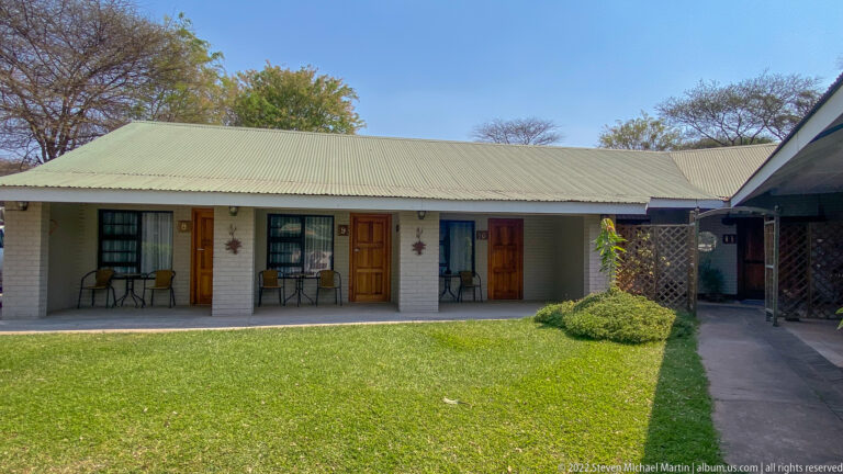 Single-story guest rooms with chairs face a green lawn beneath a corrugated roof and blue sky.