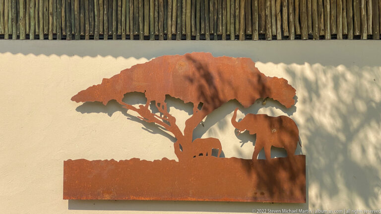 Rust-colored metal silhouette shows an elephant beneath a broad tree on a pale wall.