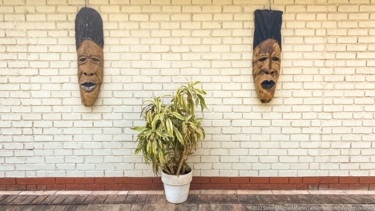 Two elongated carved wooden masks hang on a white brick wall above a potted plant.