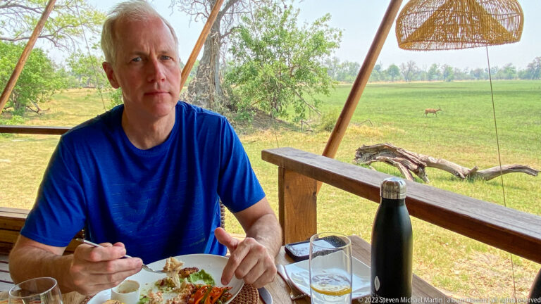 Safari guest eating lunch at an outdoor table with green floodplain and a distant antelope behind.