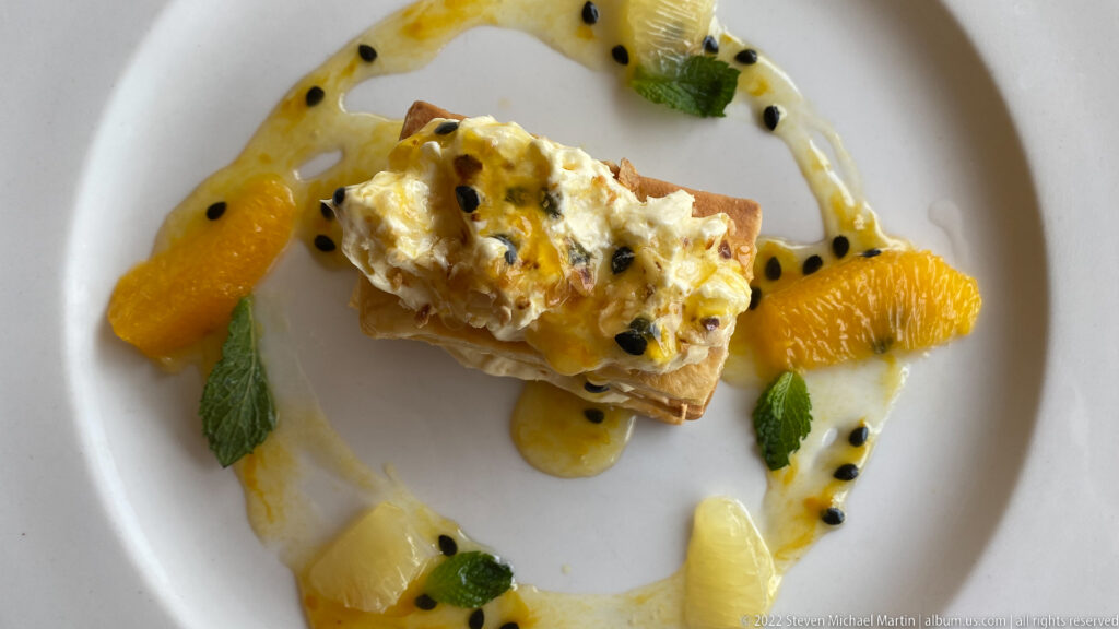 Layered pastry topped with passion-fruit cream and seeds, surrounded by citrus segments, mint, and fruit sauce.