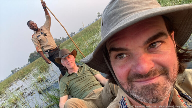 Two safari guests seated in a mokoro with a standing poler behind them in shallow wetland water.