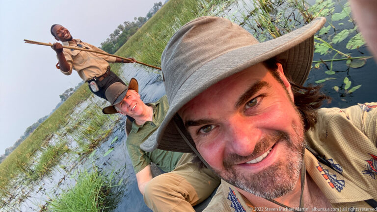 Smiling safari guest in foreground of a mokoro, with another guest and standing poler behind among water lilies.
