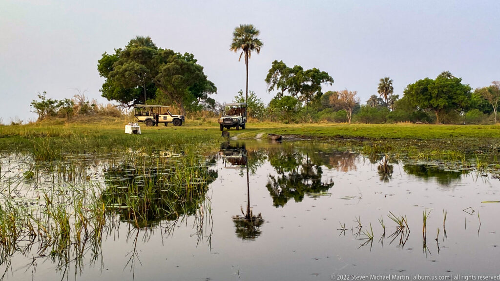 Two safari vehicles on a grassy bank beyond shallow reflective wetland water, with trees and a tall palm behind.