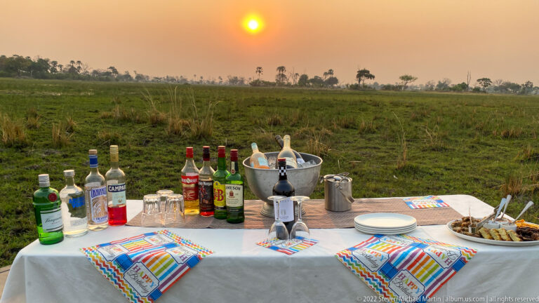 Birthday sundowner table with spirits, sparkling wine on ice, snacks, and colorful napkins beneath the setting sun.