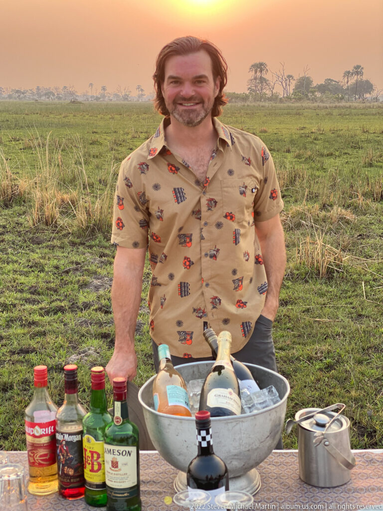 Smiling safari guest standing behind a birthday sundowner table with bottles and sparkling wine on ice.