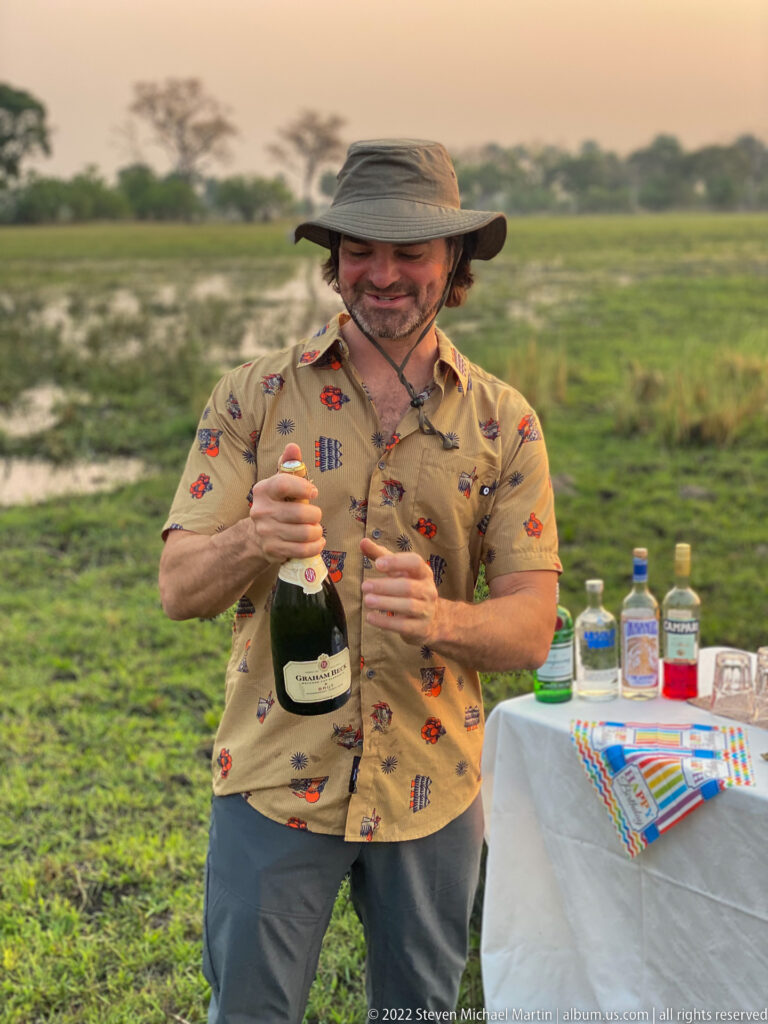 Safari guest opening a bottle of sparkling wine beside the birthday sundowner table on the floodplain.