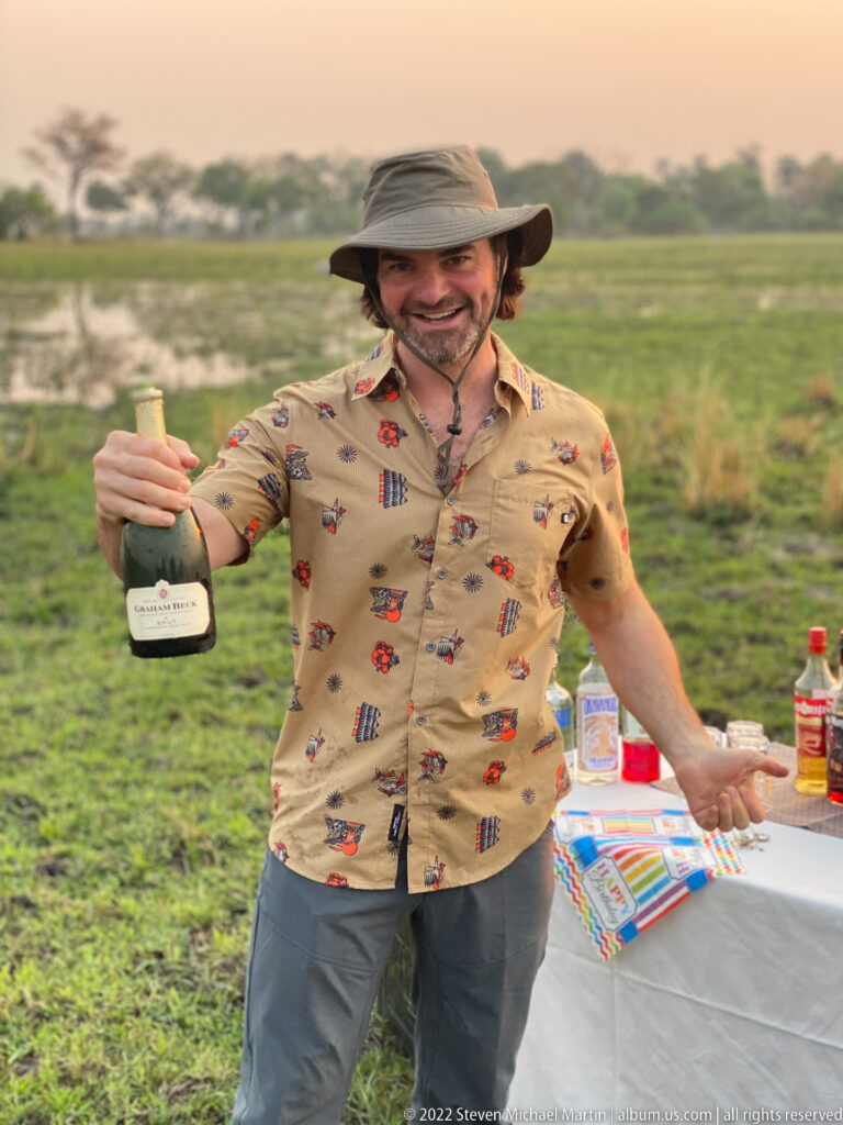 Smiling safari guest extending an opened bottle of sparkling wine beside a birthday sundowner table.