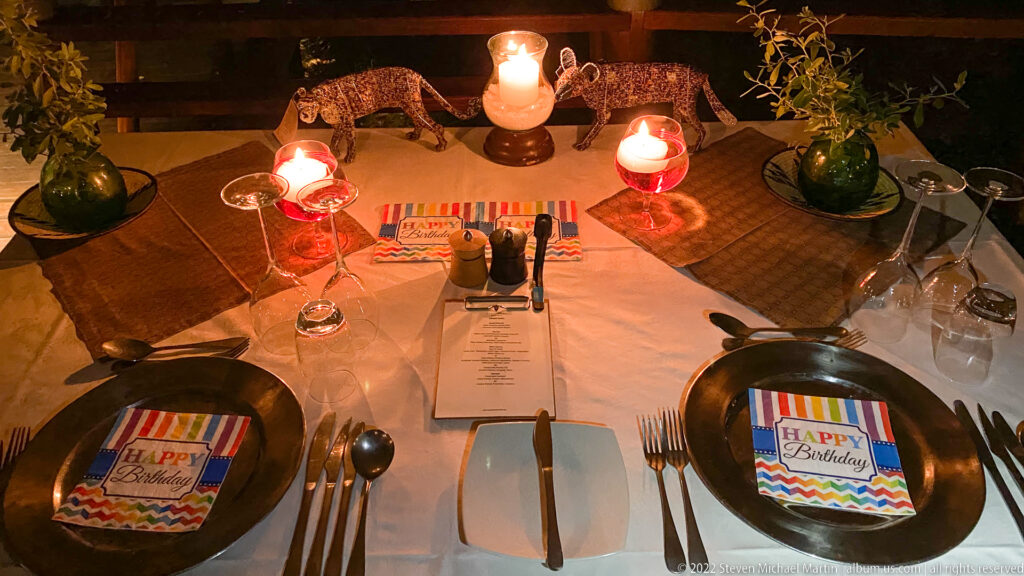 Candlelit birthday dinner table with black plates, Happy Birthday cards, flowers, glasses, cutlery, and a menu.