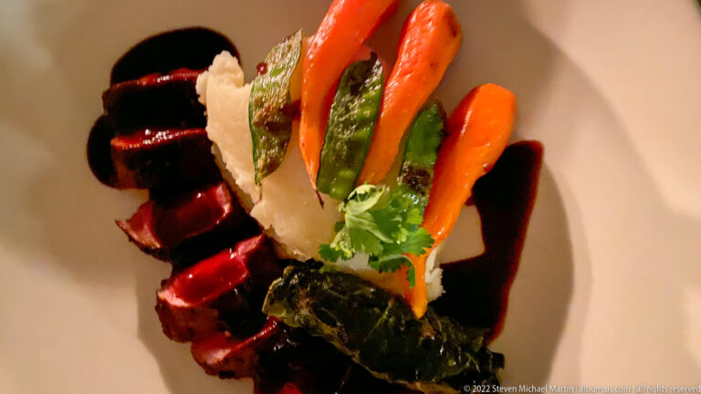 Plated vegetable course with baby carrots, asparagus, dark greens, pale crisp garnish, and glossy dark sauce.