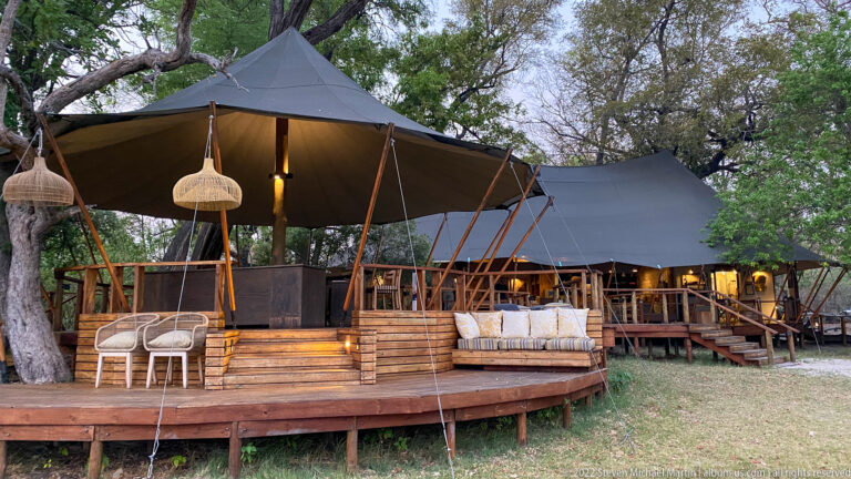Raised canvas lodge structures with timber decks, peaked roofs, woven lamps, seating, and mature trees at Stanley’s Camp.