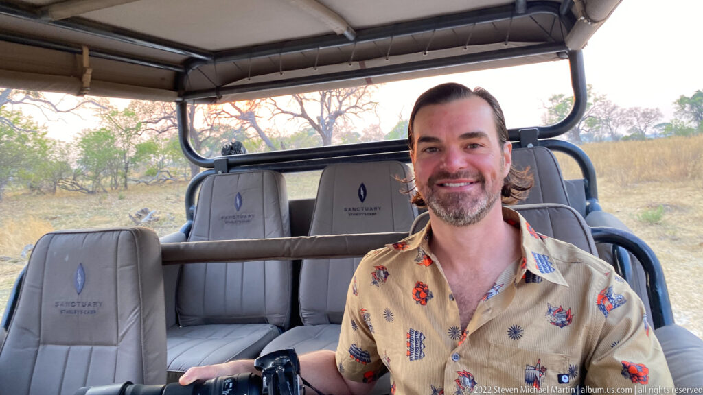 Smiling safari guest seated in an open game-drive vehicle with camera equipment and dry woodland behind.