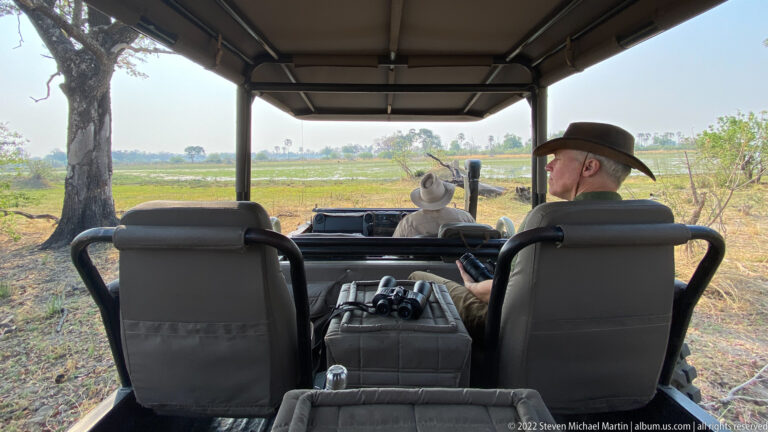 Rear-seat view inside an open safari vehicle as a guide and passenger look across a green floodplain.