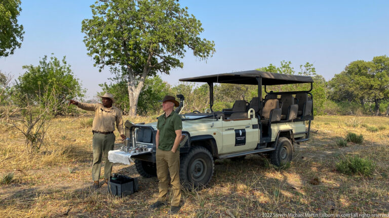 Safari guide gesturing toward the bush while a guest stands beside an open game-drive vehicle.