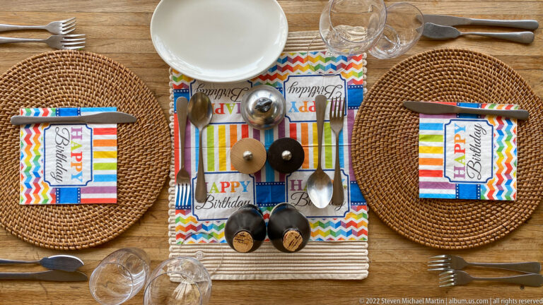 Overhead birthday table setting with rainbow napkins, Happy Birthday cards, two small cakes, candles, plates, and glassware.