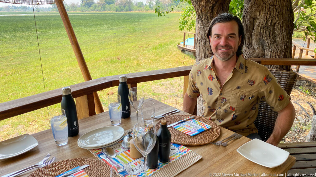 Smiling safari guest seated at a decorated birthday lunch table on a wooden deck overlooking green floodplain.