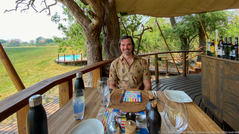Safari guest seated at a birthday table on a wooden deck overlooking open green floodplain.