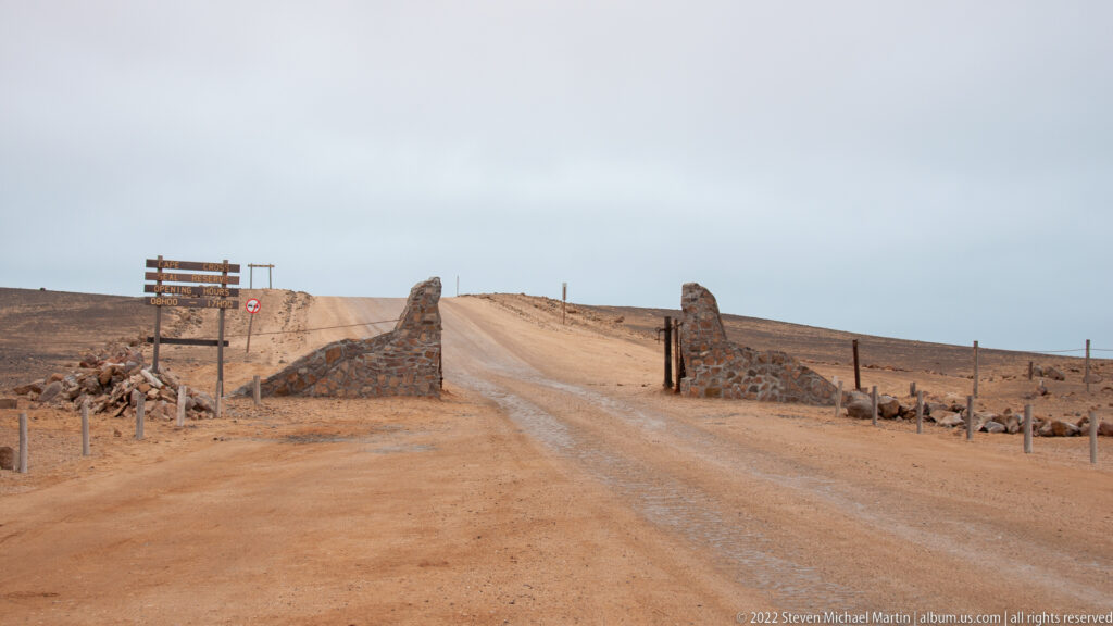 Wide view of the gravel entrance road to Cape Cross Seal Reserve in Namibia, passing between low stone gate walls in a barren coastal desert landscape.