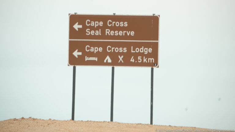 Brown road sign against a pale sky pointing to Cape Cross Seal Reserve and to Cape Cross Lodge with camping symbols, marked 4.5 km away.