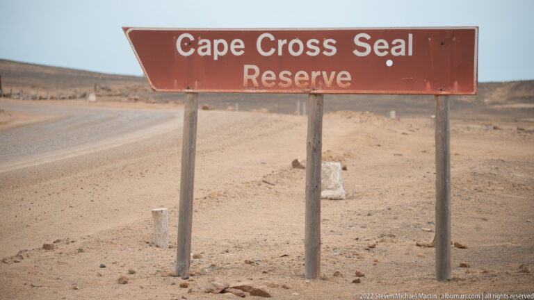 Weathered roadside sign reading “Cape Cross Seal Reserve” beside an unpaved desert road in Namibia.