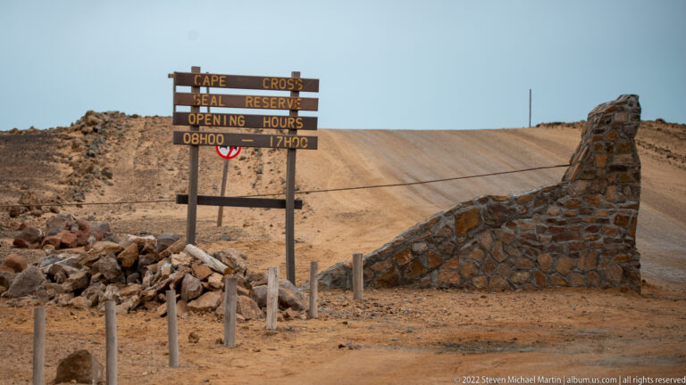 Hand-painted wooden entrance sign for Cape Cross Seal Reserve showing opening hours from 08H00 to 17H00 above a barrier across the road.