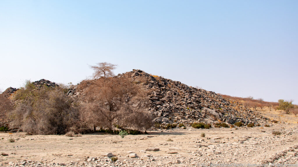 Low rocky ridge beside a pale dry wash, with thorn scrub and a sparse tree under a clear blue sky.
