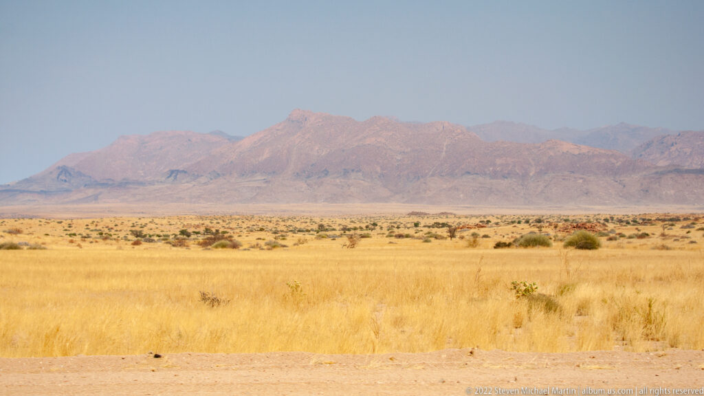 Distant hazy mountain range rising beyond a wide plain of golden dry grass and scattered shrubs.