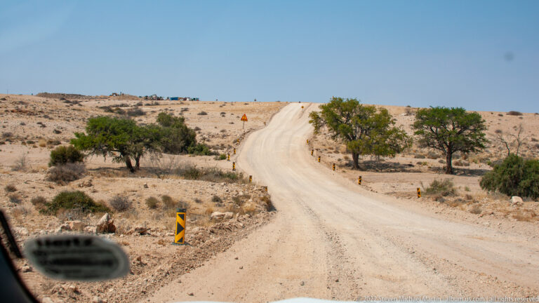 Unpaved road climbing a low rise through dry scrub, lined with black-and-yellow markers and a warning sign.