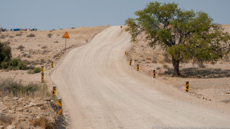 Wide gravel road curving uphill past a large tree, with striped roadside markers across pale arid ground.