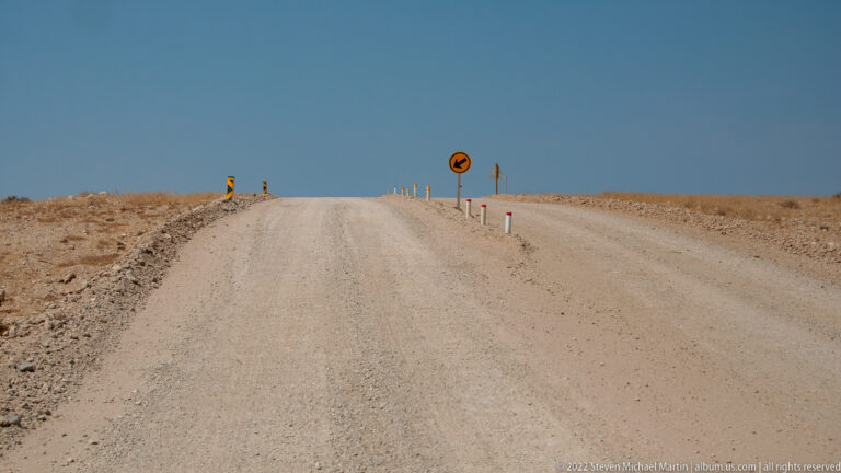 Gravel road rising to a bare horizon with a curve warning sign and sparse roadside marker posts.
