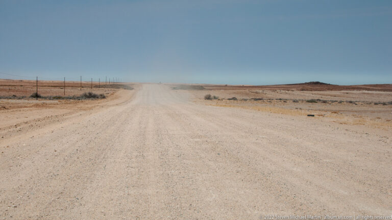 Remote gravel road curving through barren desert with a dust plume ahead and a fence line along the left.