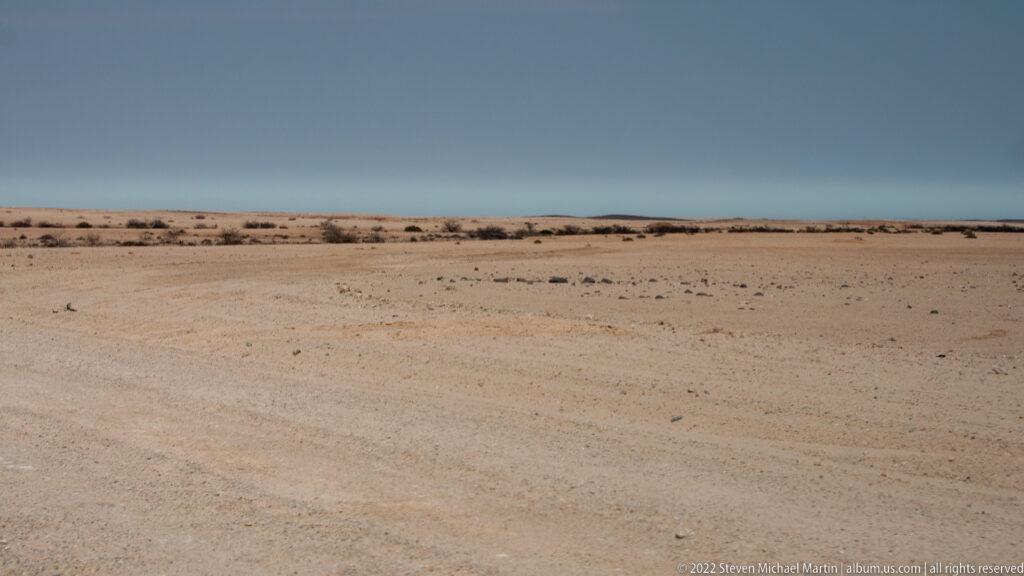 Wide pale gravel desert plain with sparse low scrub beneath a hazy blue horizon.