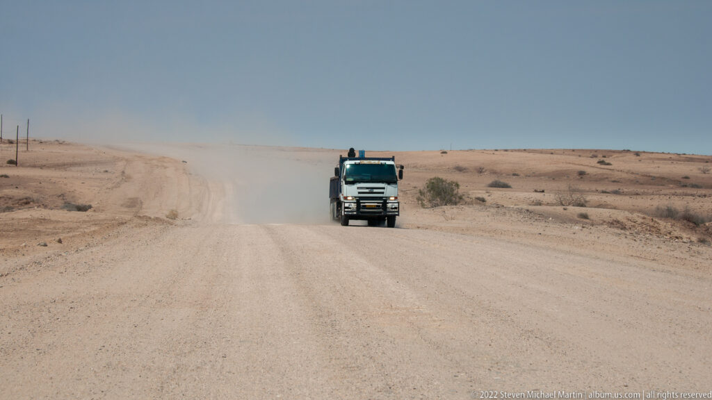 Oncoming heavy truck driving through dust on a wide gravel road across a nearly barren desert plain.