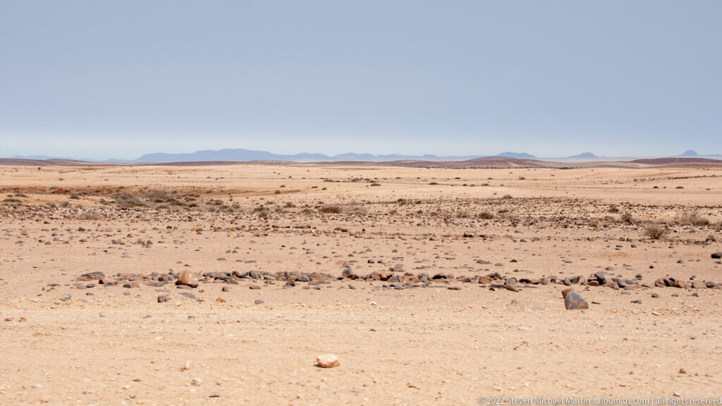 Rocky desert plain with sparse scrub extending toward low blue-gray mountains and isolated reddish hills.