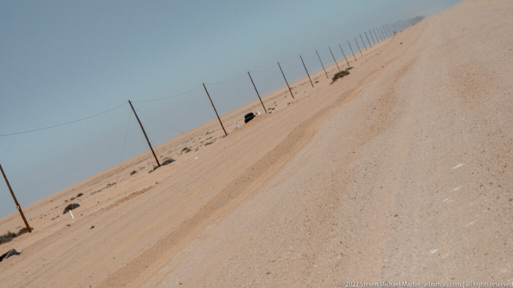 Tilted view of a pale gravel road and utility poles receding diagonally across barren desert.