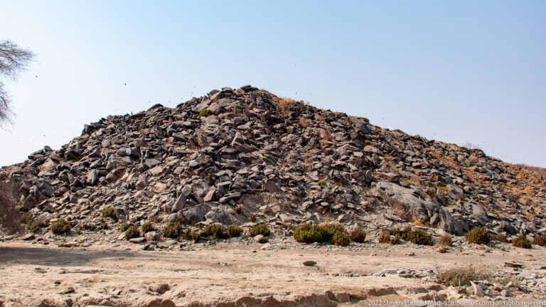 Steep mound of angular dark rocks rising from sandy ground with scattered low desert shrubs.