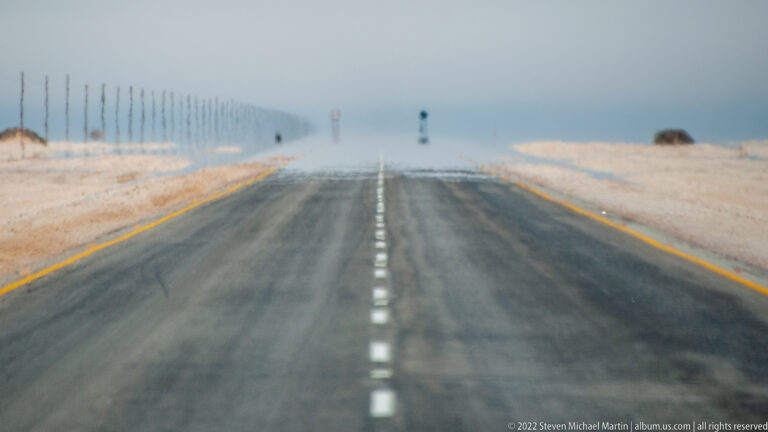 Straight paved desert highway with dashed centerline, yellow edges, and strong heat shimmer distorting the distant road.