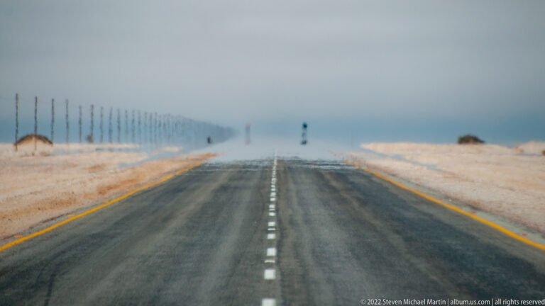 Dark asphalt highway running straight through pale desert, with distant roadside posts wavering in heat haze.