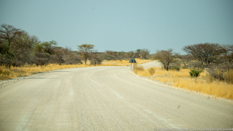 Wide gravel road curving through golden dry grass and scattered green trees, with a small roadside sign near the bend.