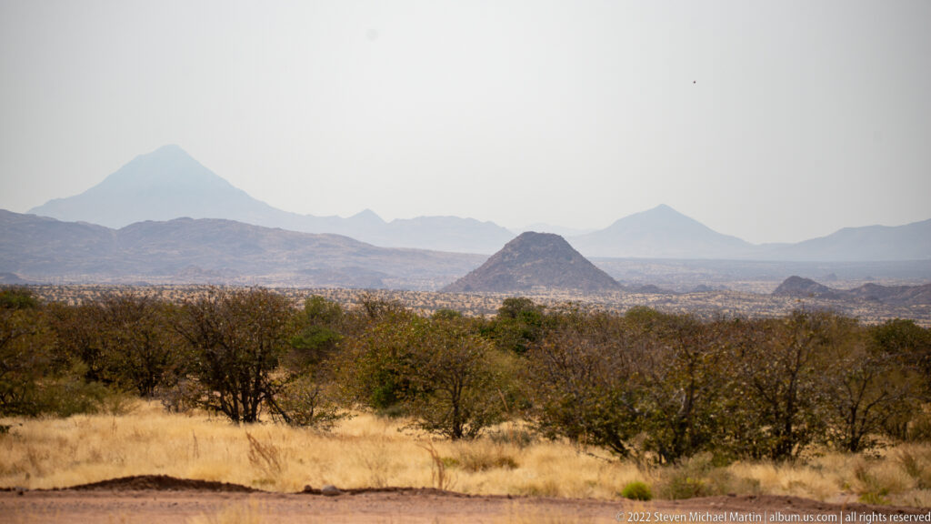 Layered rocky hills and hazy mountain peaks beyond dry grass and scrub, with a small conical hill near center.