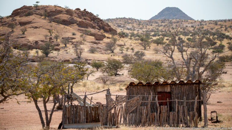 Rustic branch-built shelter and fenced enclosure beneath trees, with rocky slopes and a conical hill behind.