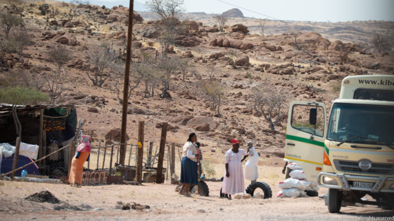 Several people beside a stopped truck, sacks, tires, and simple roadside stalls below a rocky hillside.